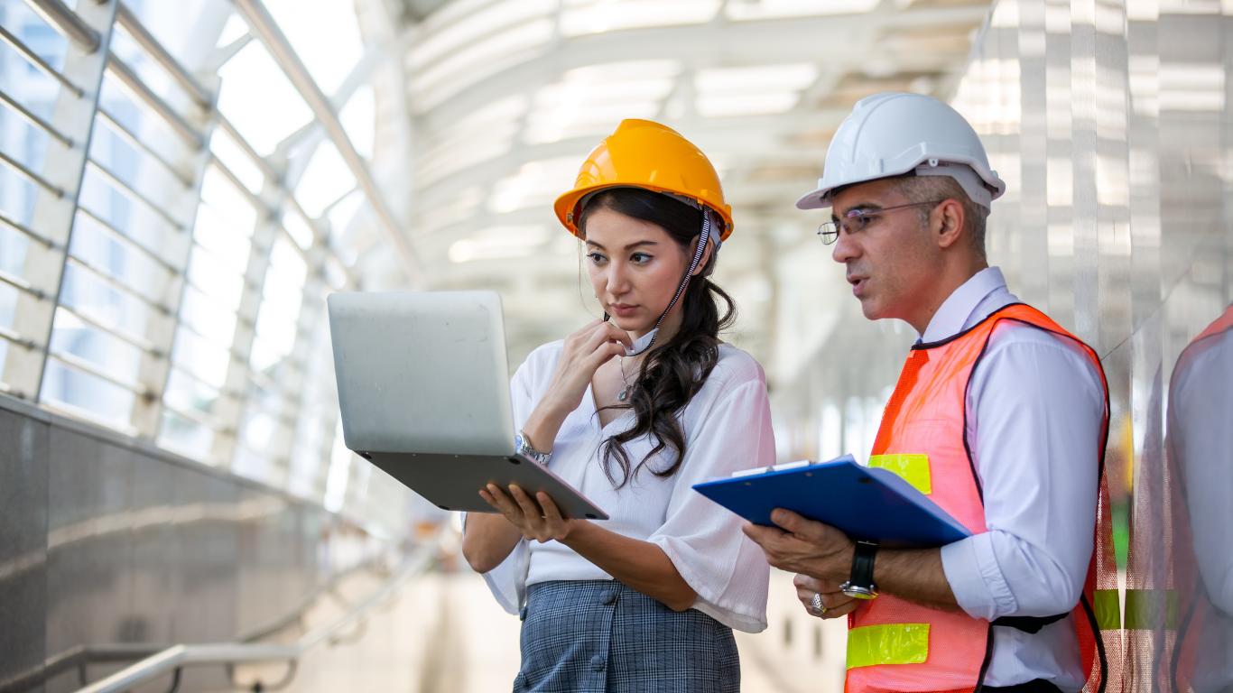 Male and female trade workers wearing construction helmets while looking at a computer